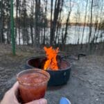 campfire with Rainy River in the background and a hand holding a Border Town Bold Caesar cocktail.