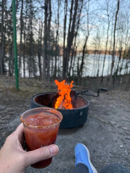 campfire with Rainy River in the background and a hand holding a Border Town Bold Caesar cocktail.